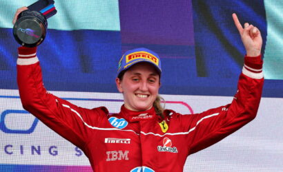 Smiling female race car driver in a red racing suit on the podium, raising a trophy in victory stance.