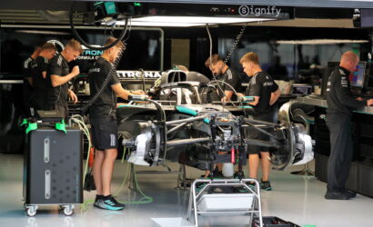 Formula 1 team members in black uniforms working around a race car on a stand in a showroom-style garage.