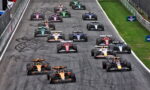 Aerial view of a Formula 1 race start with multiple cars lined up on a track, showing colorful liveries and tire skid marks.