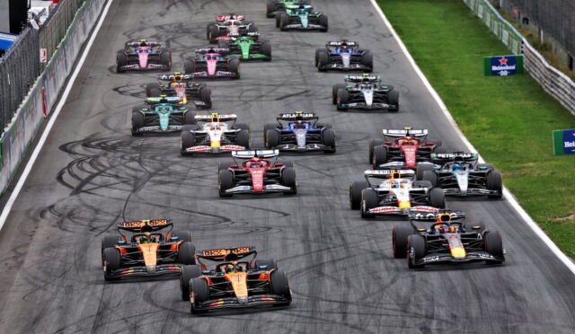 Aerial view of a Formula 1 race start with multiple cars lined up on a track, showing colorful liveries and tire skid marks.