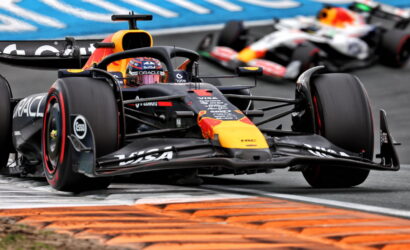 Red Bull Formula 1 car leads around a corner, with orange curbs and a blurred second car behind on the track.