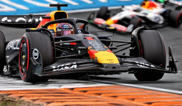Red Bull Formula 1 car leads around a corner, with orange curbs and a blurred second car behind on the track.