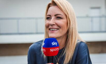 Female sports reporter smiling while holding a blue and red Sky Sports microphone outdoors.