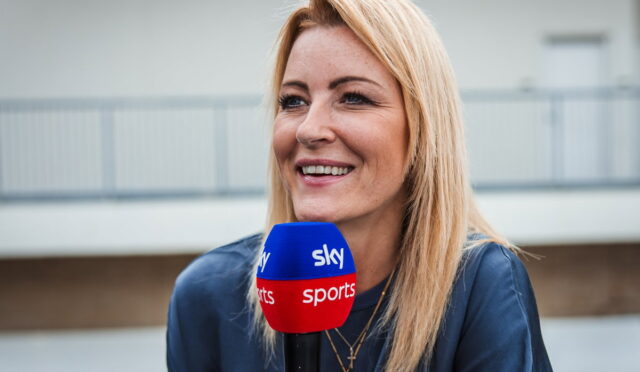 Female sports reporter smiling while holding a blue and red Sky Sports microphone outdoors.