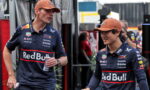 Two Red Bull Racing drivers in navy jerseys and orange caps walk and chat in a paddock area, one holding a can.
