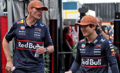 Two Red Bull Racing drivers in navy jerseys and orange caps walk and chat in a paddock area, one holding a can.