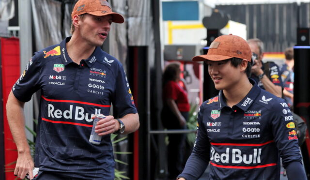 Two Red Bull Racing drivers in navy jerseys and orange caps walk and chat in a paddock area, one holding a can.