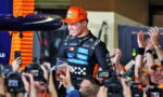 Driver in an orange cap is lifted by cheering fans, cameras flashing around them in celebration.