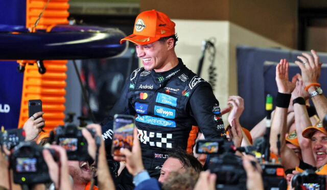 Driver in an orange cap is lifted by cheering fans, cameras flashing around them in celebration.