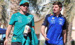 Two male Formula 1 drivers walk together outdoors, one in teal team kit holding a water bottle and jacket, the other in blue racing gear.
