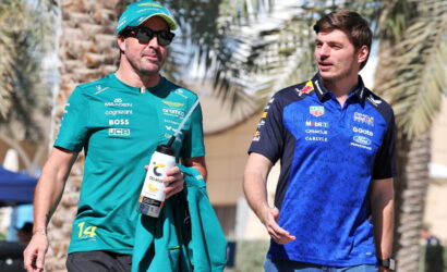 Two male Formula 1 drivers walk together outdoors, one in teal team kit holding a water bottle and jacket, the other in blue racing gear.