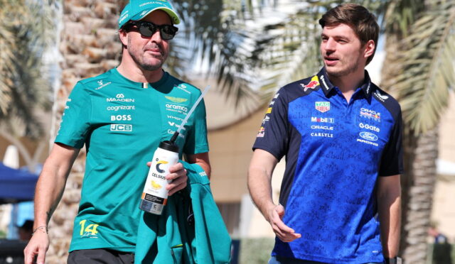 Two male Formula 1 drivers walk together outdoors, one in teal team kit holding a water bottle and jacket, the other in blue racing gear.