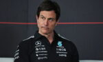 Man in a black polo shirt with race team sponsors, looking to his left against a dark backdrop.