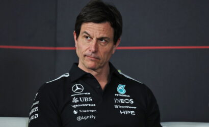 Man in a black polo shirt with race team sponsors, looking to his left against a dark backdrop.