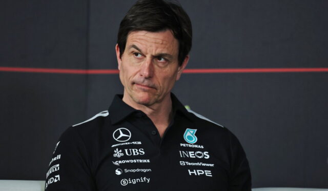 Man in a black polo shirt with race team sponsors, looking to his left against a dark backdrop.
