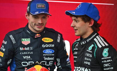 Two race car drivers in team suits stand against a red backdrop, one in Oracle Red Bull gear with a cap, the other in Mercedes gear, chatting.
