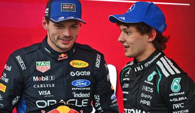 Two race car drivers in team suits stand against a red backdrop, one in Oracle Red Bull gear with a cap, the other in Mercedes gear, chatting.