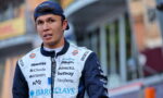 Young male race car driver in a sponsor-covered white and navy jumpsuit, standing outdoors with a backward blue cap.