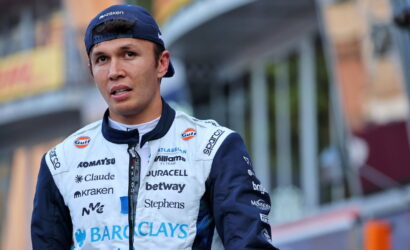 Young male race car driver in a sponsor-covered white and navy jumpsuit, standing outdoors with a backward blue cap.
