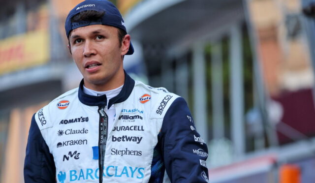 Young male race car driver in a sponsor-covered white and navy jumpsuit, standing outdoors with a backward blue cap.