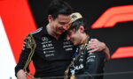 Two men on a podium share a hug, smiling, each holding large gold trophies against a bold red and black backdrop.