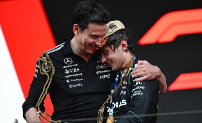 Two men on a podium share a hug, smiling, each holding large gold trophies against a bold red and black backdrop.