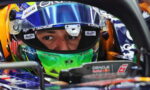 Close-up of a Formula 1 driver's face peering through a blue-green helmet visor inside a race car cockpit, focused eyes visible.