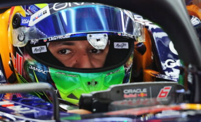 Close-up of a Formula 1 driver's face peering through a blue-green helmet visor inside a race car cockpit, focused eyes visible.