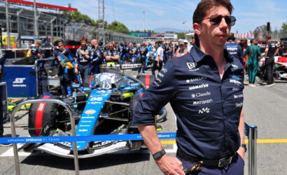 Team member in dark polo and sunglasses in the pit lane with a blue Williams F1 car behind him.