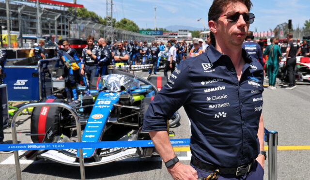 Team member in dark polo and sunglasses in the pit lane with a blue Williams F1 car behind him.
