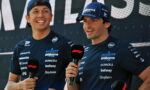 Two Williams F1 drivers in navy team shirts holding microphones during a post-race interview on stage, smiling.