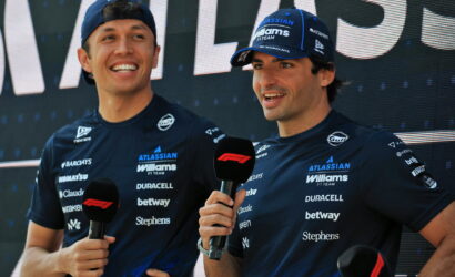 Two Williams F1 drivers in navy team shirts holding microphones during a post-race interview on stage, smiling.