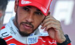 Close-up of a male race car driver in a red cap and suit, touching his ear, with visible tattoos on his hand and a beard.