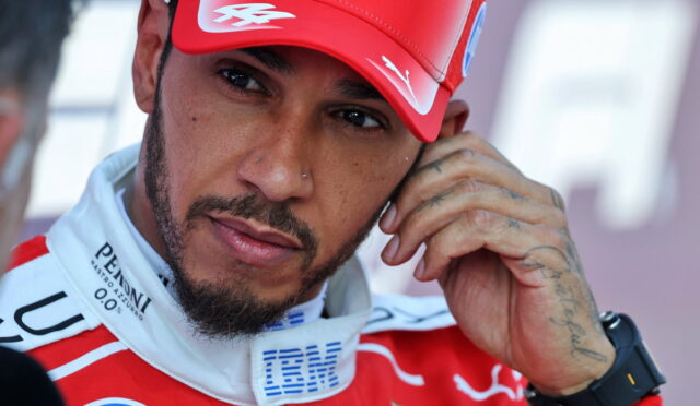 Close-up of a male race car driver in a red cap and suit, touching his ear, with visible tattoos on his hand and a beard.