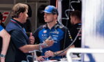 Team member talks with a race driver in blue team polo and cap in a garage/pit area, holding a drink bottle nearby.