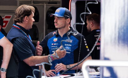 Team member talks with a race driver in blue team polo and cap in a garage/pit area, holding a drink bottle nearby.