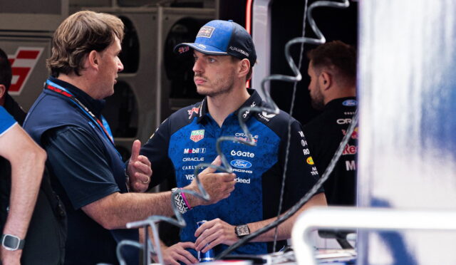 Team member talks with a race driver in blue team polo and cap in a garage/pit area, holding a drink bottle nearby.