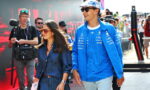 Man in blue racing jacket and cap walks beside a woman in a blue plaid dress at a busy media-covered motorsport event with photographers in the background.