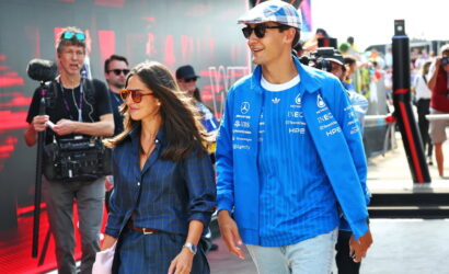 Man in blue racing jacket and cap walks beside a woman in a blue plaid dress at a busy media-covered motorsport event with photographers in the background.