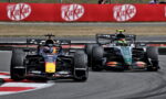Two Formula 1 cars race through a corner on a track, the Red Bull car in the lead with a dark livery and a second car close behind along the inside line, beside red advertising banners.