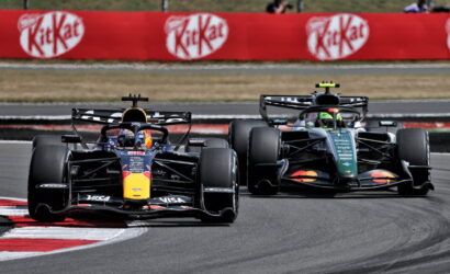 Two Formula 1 cars race through a corner on a track, the Red Bull car in the lead with a dark livery and a second car close behind along the inside line, beside red advertising banners.