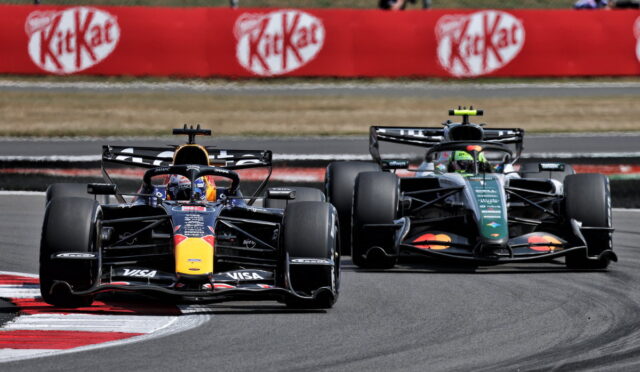 Two Formula 1 cars race through a corner on a track, the Red Bull car in the lead with a dark livery and a second car close behind along the inside line, beside red advertising banners.