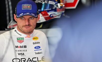 Close-up of a race car driver wearing a blue Oracle Red Bull Racing cap and a white sponsor-laden racing suit, looking ahead.