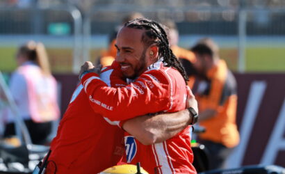 Two race car drivers in red suits share a celebratory hug on the track, smiling at each other.