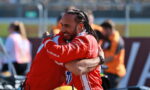 Two race car drivers in red suits hug in celebration on a track.