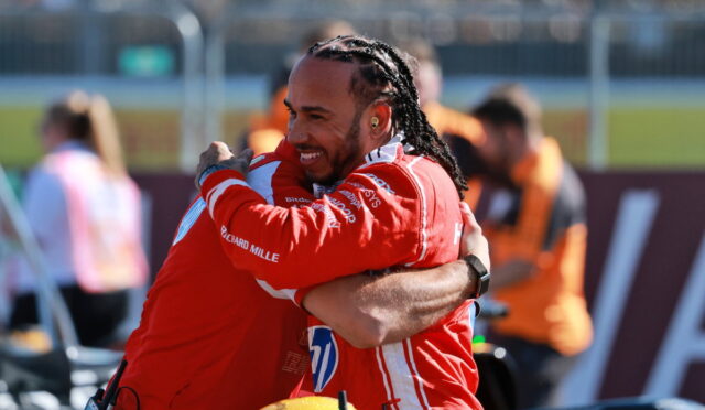 Two race car drivers in red suits hug in celebration on a track.