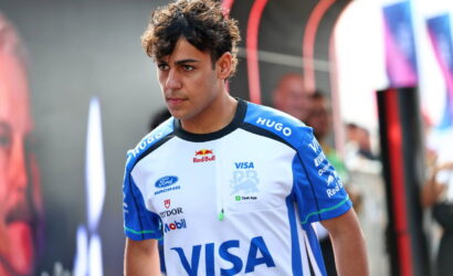 Young man in blue-and-white racing jersey with VISA logo stands in a pit area, focused ahead, sponsor logos visible on his shirt.