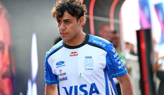 Young man in blue-and-white racing jersey with VISA logo stands in a pit area, focused ahead, sponsor logos visible on his shirt.