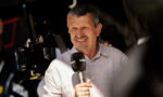 Male presenter holding a Canal+ microphone in front of camera gear during a live interview, wearing a light button-up shirt and squinting slightly in bright light.