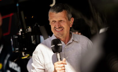 Male presenter holding a Canal+ microphone in front of camera gear during a live interview, wearing a light button-up shirt and squinting slightly in bright light.